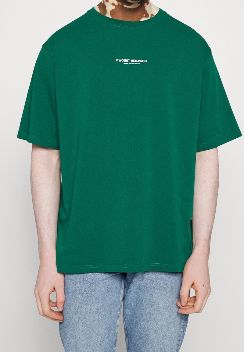 Grön bomullströja med avspänd passform, rundad hals och korta ärmar. Har vit text på framsidan som säger "VÄRSTA BETEENDE."