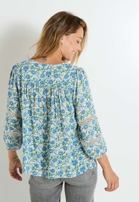 Blouse florale en tissu léger avec des motifs de fleurs bleues et vertes, un détail froncé à l'arrière et des accents en dentelle sur les manches.