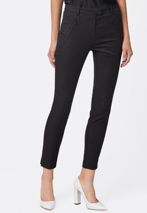 Femme portant un pantalon noir slim aux chevilles avec poches avant zippées et chaussures blanches à talons hauts, sur un fond uni.