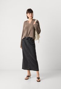Pull en maille marron avec un col en V et des détails aux épaules, associé à une longue jupe en denim noir et des sandales à talons marron. Accessoire : un simple sac à dos.