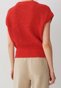 Czerwony, dziany sweter z krótkim rękawem i ściągaczem u dołu. Materiał ma grubą, teksturowaną fakturę. Połączony z beżowymi spodniami z tylnymi kieszeniami.