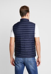 Marinblå puffer väst med horisontell quiltning, hög krage och dragkedjestängning. Buren över en vit skjorta och parat med blå jeans.