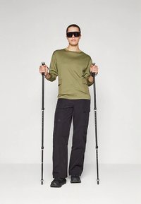 Persona che indossa una camicia a maniche lunghe verde, pantaloni cargo neri e scarpe nere, mentre tiene dei bastoni da trekking, in piedi su uno sfondo uniforme chiaro.