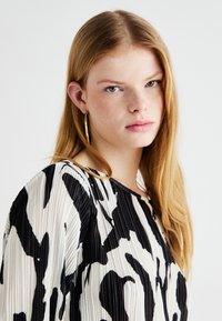 Melna un balta plīsēta blūze ar abstraktiem rakstiem, apaļa kakla izgriezuma un garām piedurknēm. Modelis valkā lielas uztveramu auskarus.