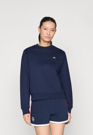 Marineblå hettegenser med ribbestrikkede mansjetter og hem, med en liten grønn logo. Kombinert med marineblå shorts med hvit kant og et emblem.