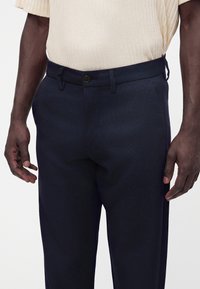 Homme portant un pantalon bleu marine foncé sur mesure et une chemise beige clair côtelée à manches courtes, debout avec les mains détendues le long du corps.