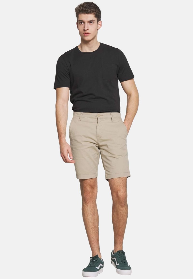 Șorturi bej deschis cu mansete răsfirate, asociate cu un tricou negru mulat și adidași verzi. Tricoul are o mică buzunăraș pe piept.