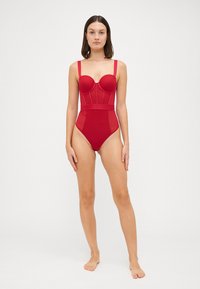 Roter Einteiler in Badeanzugform mit strukturierter Oberlinie, gepolsterten Körbchen, verstellbaren Trägern und hochgeschnittenem Beinschnitt mit durchsichtigen Einsätzen.
