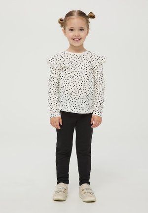 Camicia bianca a maniche lunghe con pois neri e maniche arricciate, abbinata a leggings neri e scarpe di colore chiaro.