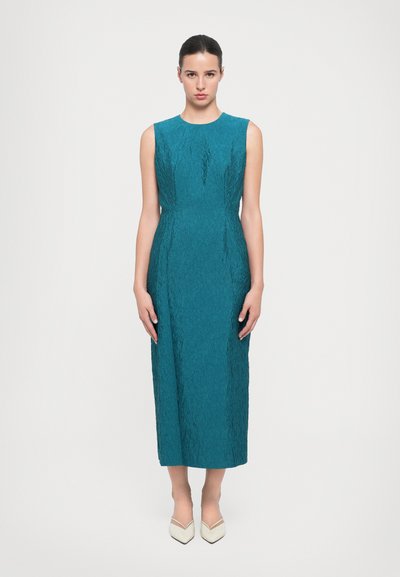 Teal sans lengan dengan pola berpola, menampilkan pinggang yang pas dan rok midi lurus. Dipadukan dengan sepatu krim berbentuk runcing.