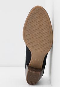 Semelle de chaussure noire avec grip en caoutchouc marron clair, talon bloc marqué "ANTI GLISSE", taille 37, inclinée contre un mur blanc et un sol blanc.
