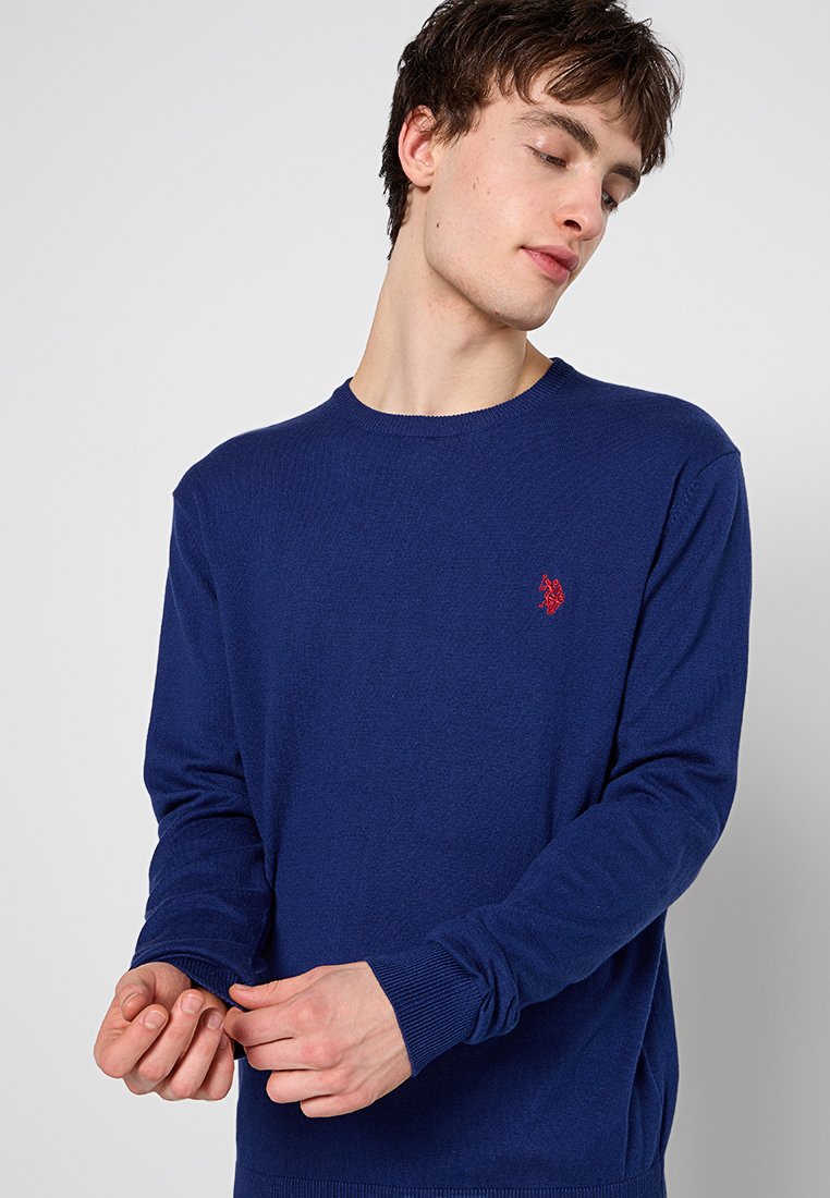 Navyblauer Strickpullover mit Rundhalsausschnitt, langen Ärmeln, gerippten Bündchen und einem kleinen roten Emblem auf der Brust. Weiche Textur, mittleres Gewicht.