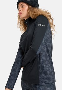 Junge Frau in einer schwarzmusterigen Roxy Reißverschlussjacke und Strickmütze, die mit gebogenen Armen vor einem weißen Hintergrund posiert.