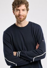 Pull en tricot bleu marine avec un col rond côtelé, des surpiqûres blanches sur les manches et des détails de boutons à l'épaule. Texture douce.