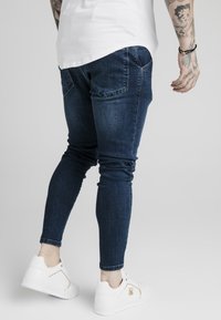 Jeans ajustados de mezclilla azul oscuro, con bolsillos traseros y un leve estiramiento. Combinados con zapatillas blancas con detalles dorados.