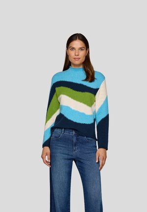 Frau trägt einen mehrfarbigen gestreiften Pullover in Blau, Grün, Weiß und Marineblau mit blauen Jeans und steht vor einem schlichten grauen Hintergrund.