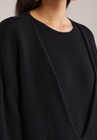 Juodas, tekstūruotas megztinis su plačiu apykakle ir nusileidusių pečių siūlėmis, demonstruojantis minkštą, jaukų mezginį.