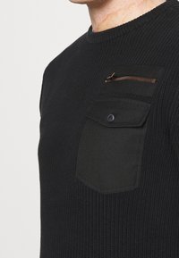 Schwarzer Strickpullover mit rundem Ausschnitt, ausgestattet mit einer Brusttasche mit einem bronzefarbenen Reißverschluss und rechteckigem Klappenverschluss, strukturierte Textur.