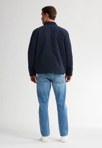 Marineblauwe jas met een ontspannen pasvorm, gecombineerd met lichtblauwe jeans. De outfit wordt compleet gemaakt met witte sneakers. Zichtbare schoudernaden en knopen.