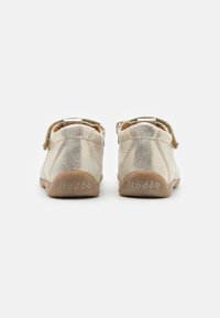 Un par de zapatos para niño pequeños en beige con una correa de velcro, costuras visibles y suelas flexibles marrones de la marca "froddo", mostrados desde la parte trasera.