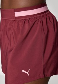 Burgundinfärgade träningsshorts med ett elastiskt midjeband i rosa och vitt samt en liten vit Puma-logotyp på vänster sida. Lättviktigt material.