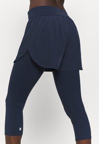 Námornické sportovní šortky přes odpovídající capri legíny, vyrobené z lehkého materiálu. Šortky mají perforované detaily podél lemu.