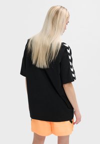 Zwart oversized T-shirt met korte mouwen, voorzien van een geometrisch wit patroon op de schouders. Gecombineerd met oranje shorts.
