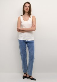 Top in cotone bianco a coste con orlo scallop, abbinato a jeans blu chiaro a gamba dritta e mule nere con tacco. La modella è in piedi di fronte a uno sfondo neutro.
