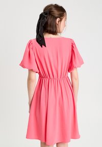 Robe rose à manches courtes avec une taille cintrée et une jupe évasée. Présente un détail au niveau du col au dos et un ruban noir dans les cheveux. Texture de tissu lisse.