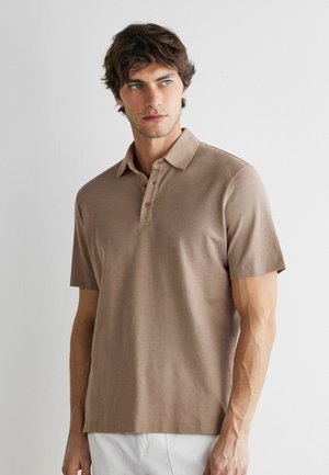 Mężczyzna w brązowej koszulce polo z krótkim rękawem i białych spodniach, stojący na białym tle, patrzący na bok.