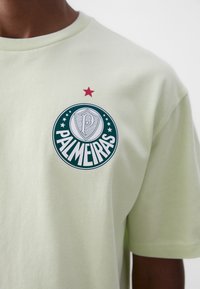 Camiseta de algodón verde claro con un logotipo redondo de Palmeiras, detalles en blanco y verde, y una estrella roja encima del logotipo, de manga corta.