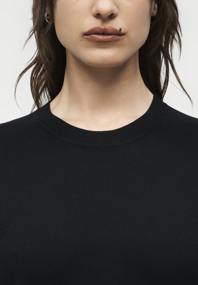 Κοντινό πλάνο του κάτω μέρους του προσώπου και του λαιμού μιας γυναίκας, που φοράει μαύρη μπλούζα με στρογγυλή λαιμόκοψη, με χαλαρά καστανά μαλλιά και ένας σπίλος κοντά στο πάνω χείλος της.