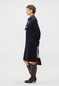 Γυναίκα με μινι φόρεμα πλέξης σε ναυτικό μπλε χρώμα, σχέδιο μαντήλι, καφέ μπότες μέχρι το γόνατο και κρατώντας καφέ τσάντα πίσω από την πλάτη της.