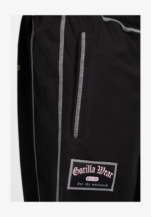 Zwarte sportbroek van een gestructureerde stof, met zilveren stiksel, een zijzak en een opvallende 'Gorilla Wear' logo patch op de dij.