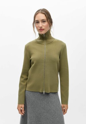 Femme portant un pull à col montant zippé vert olive et une jupe en laine grise, debout devant un fond blanc uni.