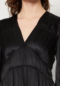 Robe noire avec décolleté en V, manches longues et plis texturés sur tout le buste, en tissu lisse avec une jupe fluide.