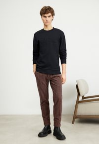 Fekete pulóver, dombornyomott logóval, barna kockás, tapered nadrággal párosítva, és fekete, vastag sarkú bokacsizmával. Minimalista dizájn, laza szabás.