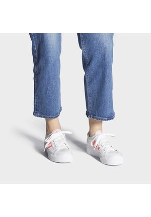 Persoon draagt korte blauwe spijkerbroek en witte sneakers met roze strepen, staand op een effen lichte achtergrond.