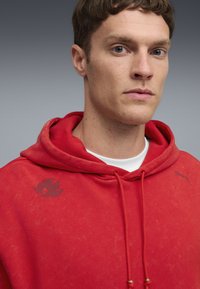 Rode katoenen hoodie met een kangoeroe-pocket, verstelbare trekkoorden en logo's op de borst. Zachte textuur met een licht vervaagd effect.