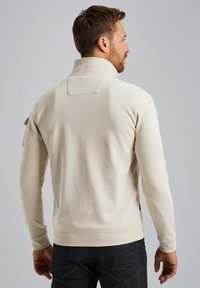 Sweatshirt boje kreme s visokim ovratnikom, elegantnim dizajnom i rebrastim manžetama. Sadrži izrez na leđima i bočnu džep.