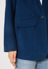 Tummansininen corduroy-takki, jossa on pystyribbaus, yksittäinen napitus ja vasemmassa rintataskussa. Pehmeä rakenne; rento istuvuus.