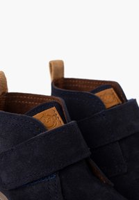 Scarpe scure in suede blu con un accento in pelle marrone e cinturino in velcro. Dettagli di cucitura evidenti lungo i bordi e soletta con motivo.