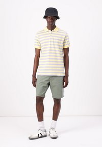 Polo à manches courtes jaune avec des rayures lavande, associé à un short vert. Chapeau seau noir et baskets blanches avec des accents noirs.