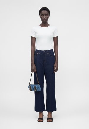 Femeie care poartă un top alb cu mâneci scurte și blugi largi albastru închis, ținând o geantă mică albastră, stând în fața unui fundal simplu.