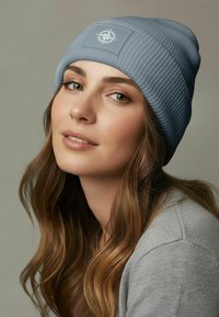 Sky blue ribbed beanie with a compass logo patch; soft texture; worn with a gray sweater; medium-length wavy hair framing the face.