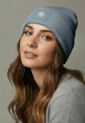 Sky blue ribbed beanie with a compass logo patch; soft texture; worn with a gray sweater; medium-length wavy hair framing the face.