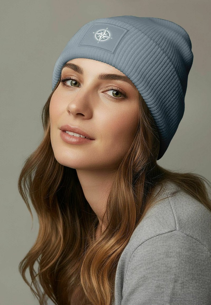 Sky blue ribbed beanie with a compass logo patch; soft texture; worn with a gray sweater; medium-length wavy hair framing the face.