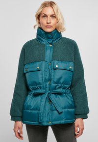 Veste matelassée teal avec un haut en polaire texturé. Elle présente un col haut, des boutons de fermeture, deux poches avant et une taille ceinturée.