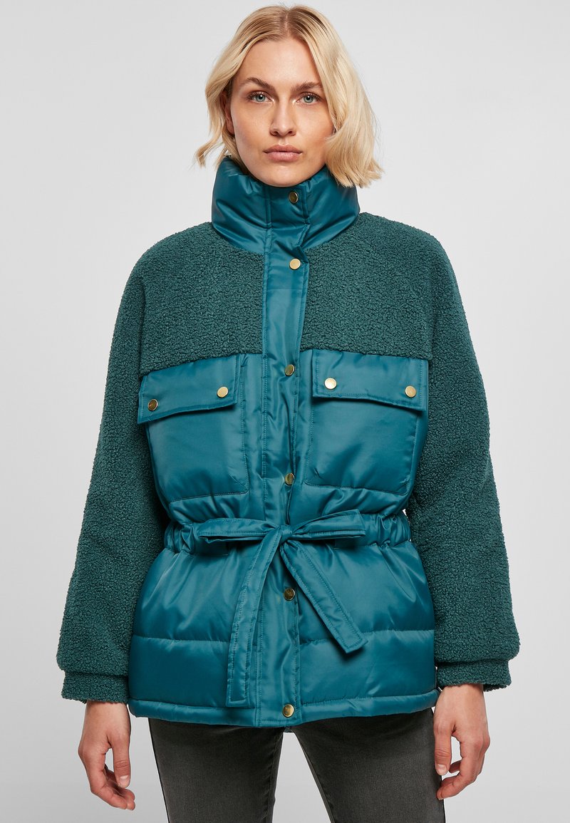 Veste matelassée teal avec un haut en polaire texturé. Elle présente un col haut, des boutons de fermeture, deux poches avant et une taille ceinturée.