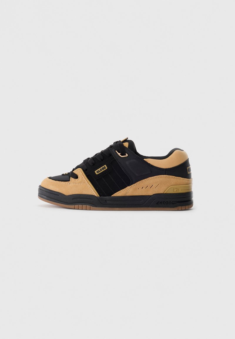 Basket de skate basse en suède noir et tan avec des lacets noirs, étiquette de marque sur le côté et semelle en caoutchouc à motif, présentée de côté sur un fond blanc.